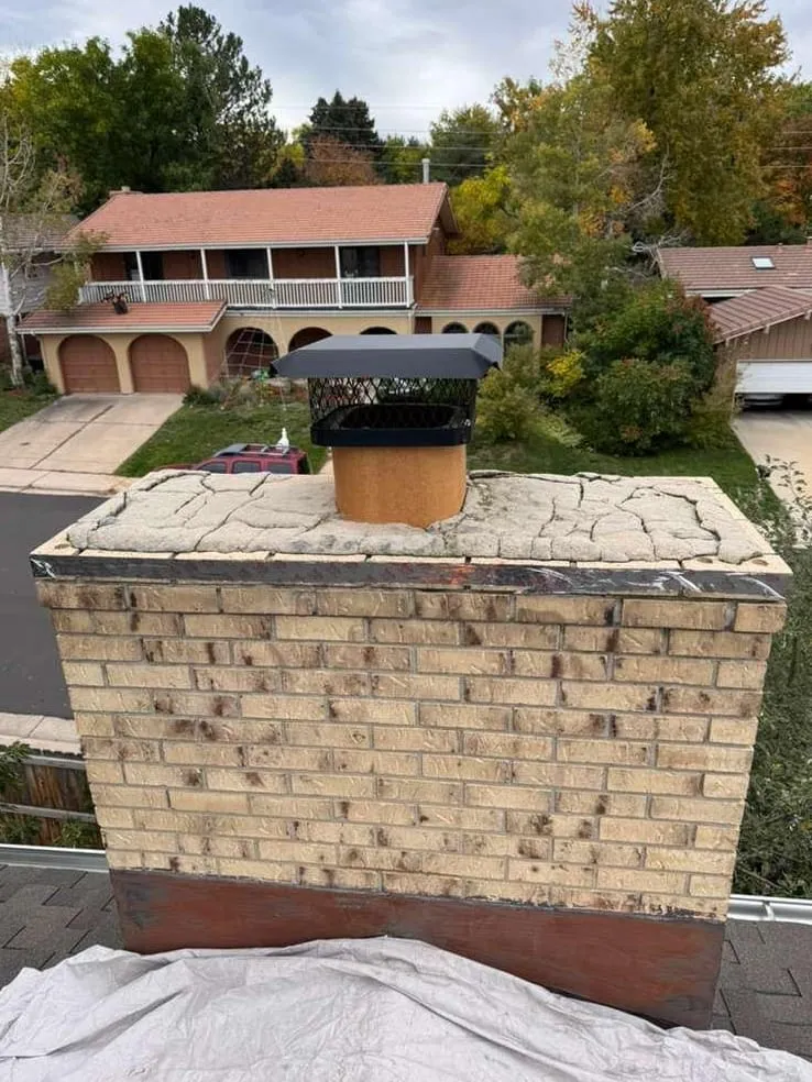 A buff brick chimney with a crazed, cracked concrete crown, a black mesh cap on an orange collar and a rust-brown band at its foot, with a grey tarp on the roof below