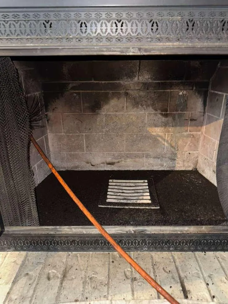 A sooty firebox under a decorative metal frame, with a cleared floor and a white ash grate, an orange rod laid across it and a black mesh curtain at the left