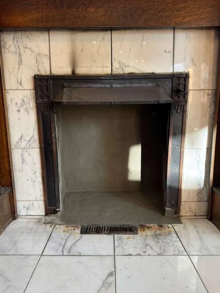 A clean, empty firebox in a white marble hearth with a dark iron insert frame and an oak mantel above, a small floor grille below