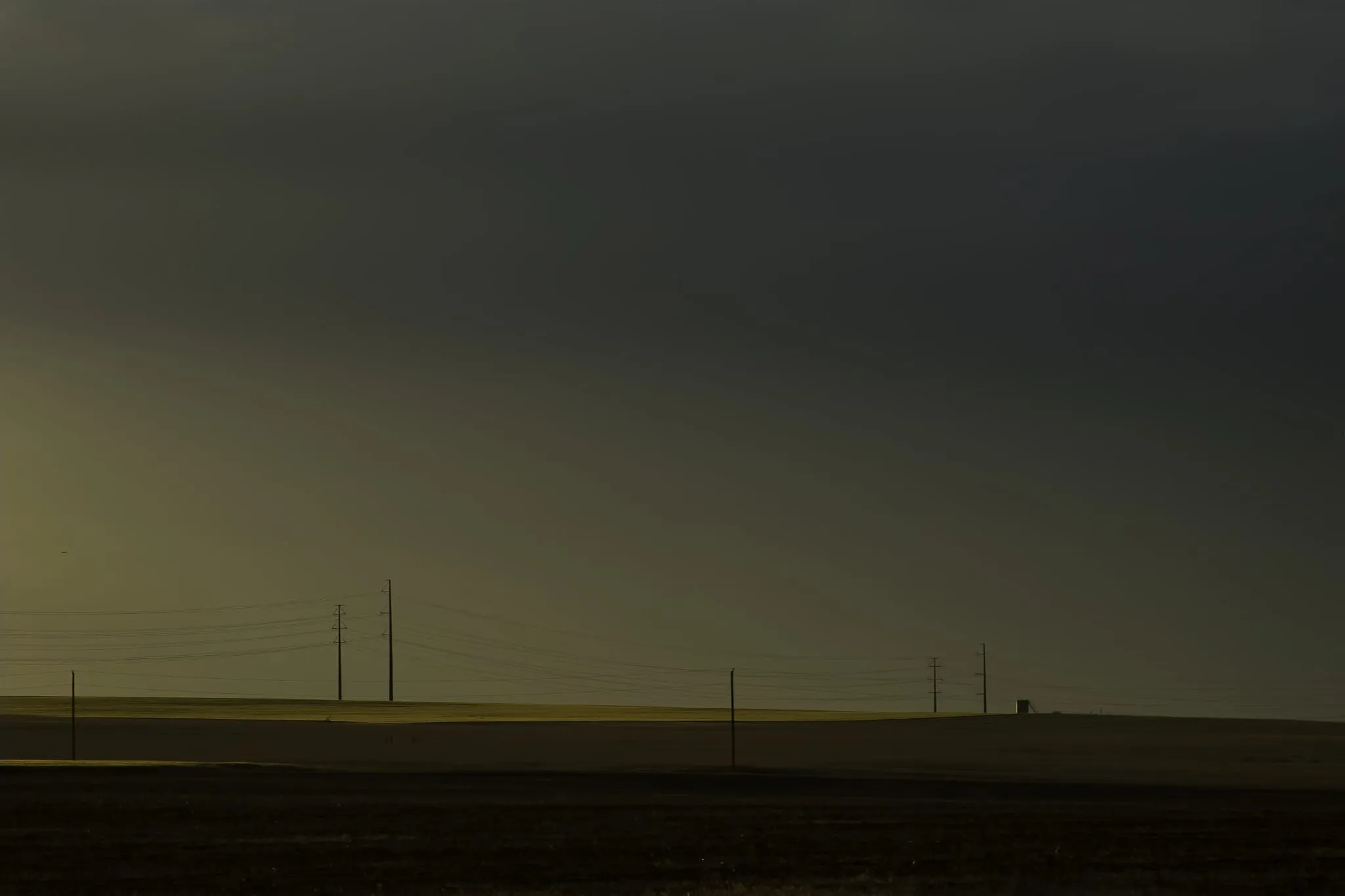 A dark, heavy sky over flat farmland at dusk, with a line of utility poles and wires along the horizon and a strip of light across the field